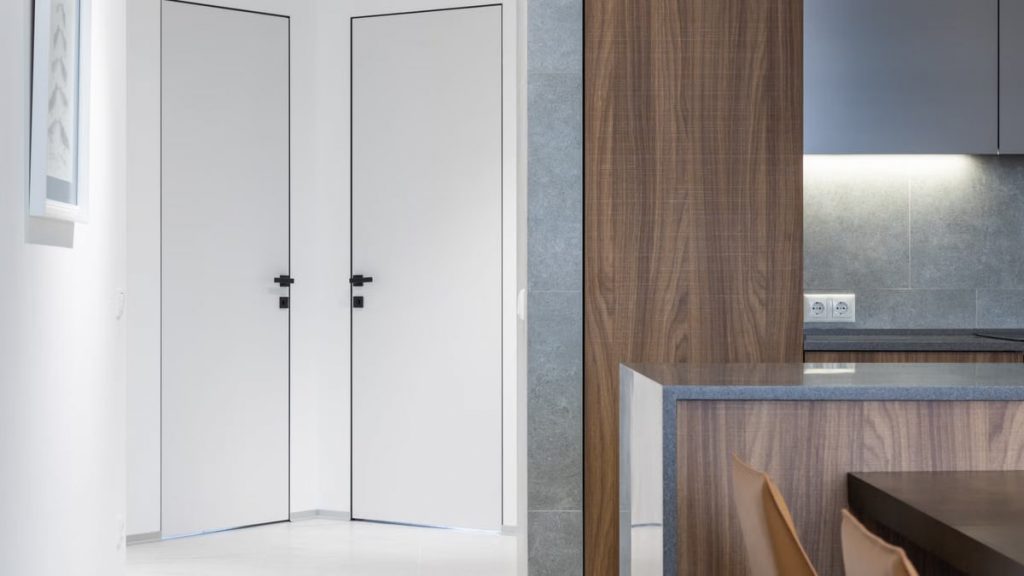 Hallway with white doors and walls near wooden kitchen counter with cupboards near table and chairs in modern apartment at home